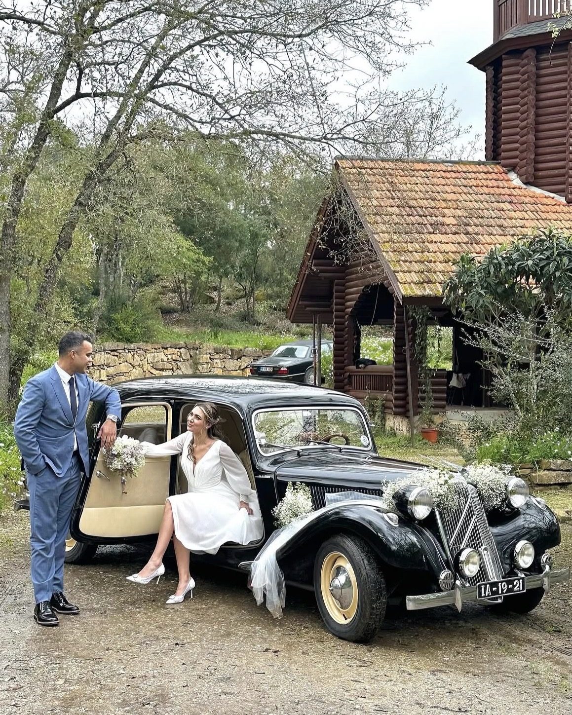 Noiva a sair de Citroën Traction preto decorado com flores, junto a noivo de fato azul, A&M Wedding Cars